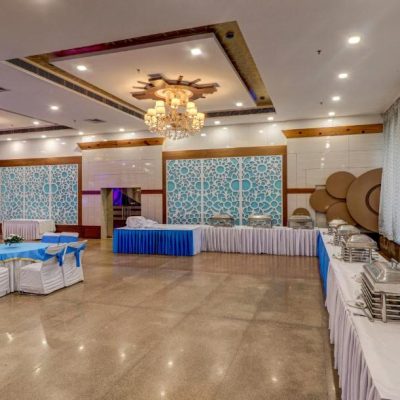 A decorated banquet hall with round tables and chairs covered in white and blue cloth, chandeliers on the ceiling, buffet tables with covered dishes, and patterned blue decorative panels on the walls.